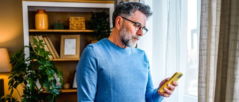 A middle-aged man with a beard and glasses, wearing a light blue sweater and mustard yellow pants, stands by a window and looks intently at a yellow smartphone he holds in his hand.